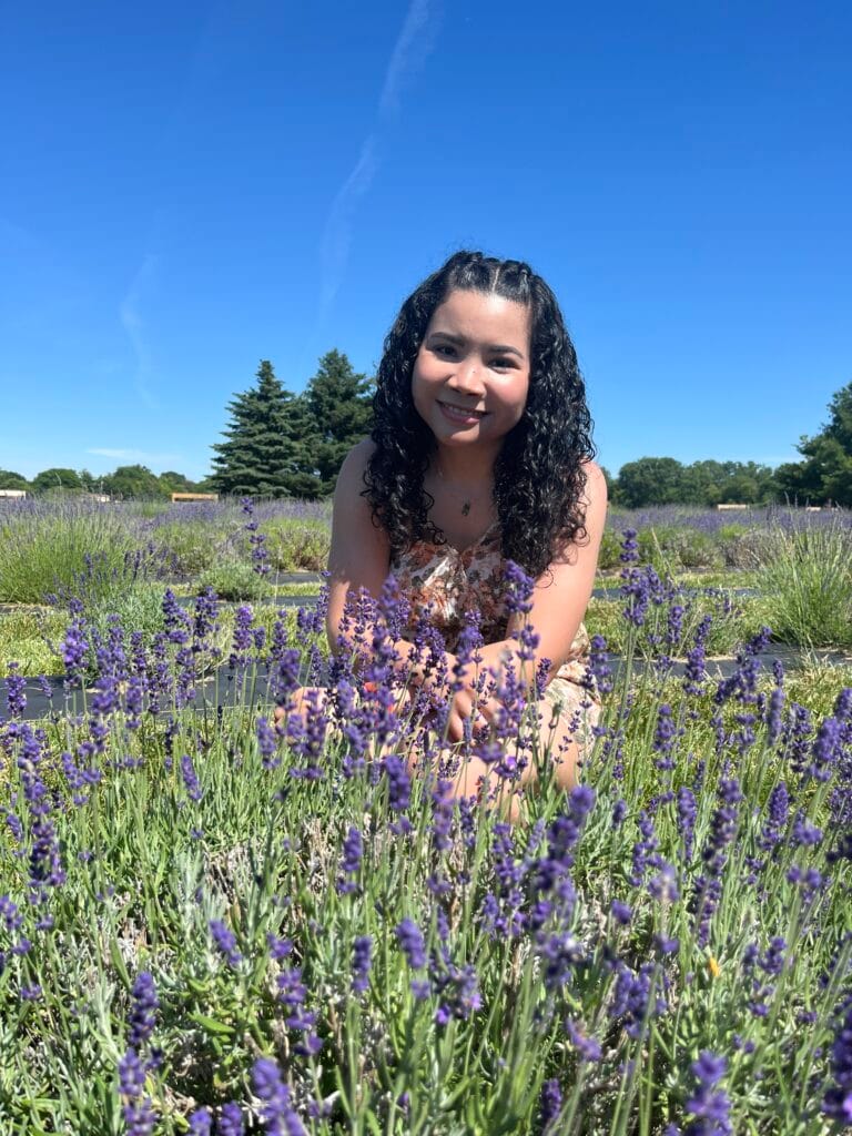Angélica Madrid sonriendo entre flores de lavanda en un campo bajo un cielo azul.