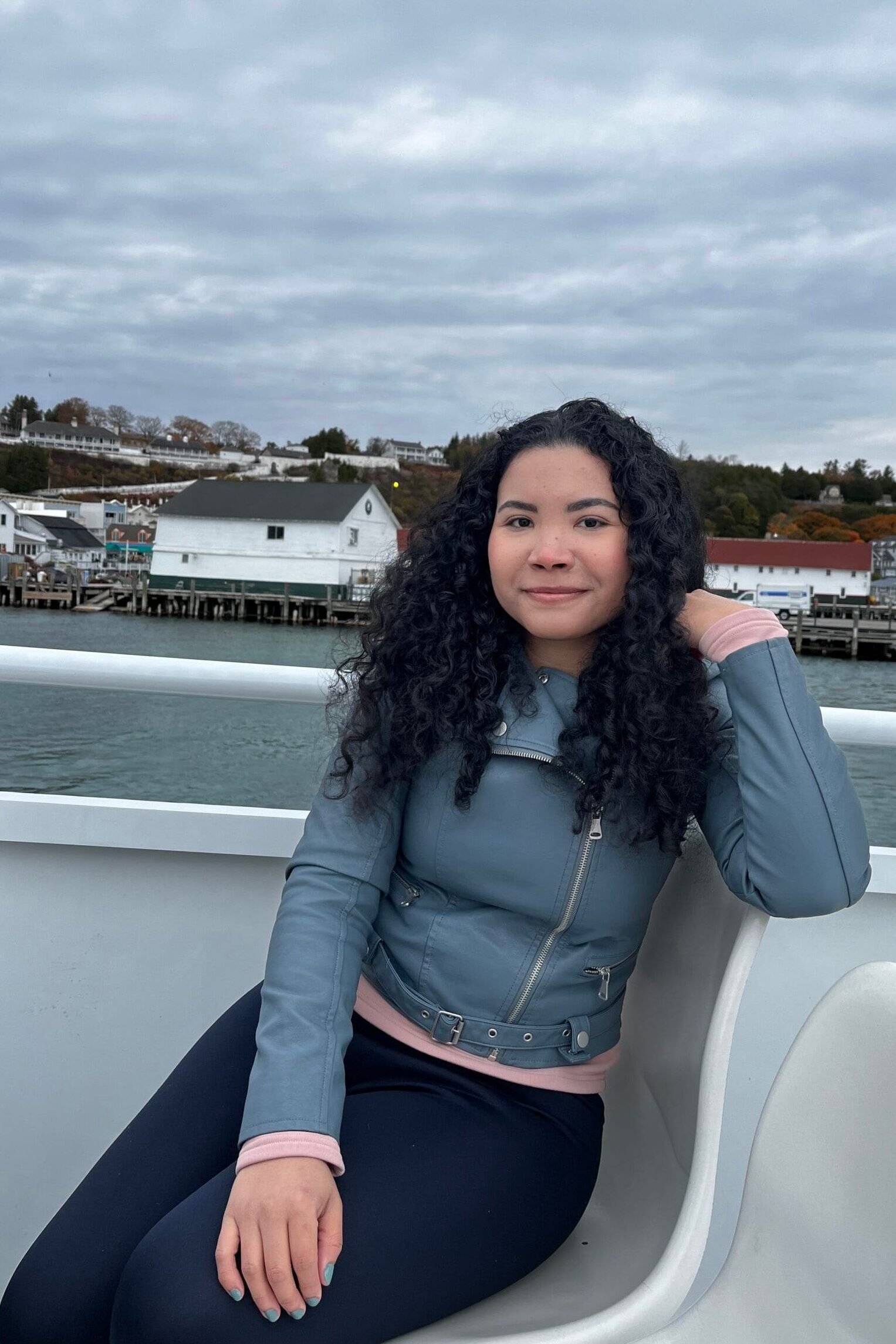 Angélica Madrid sentada en un barco, con fondo de casas junto al agua y cielo nublado, durante un viaje por Mackinac Island.